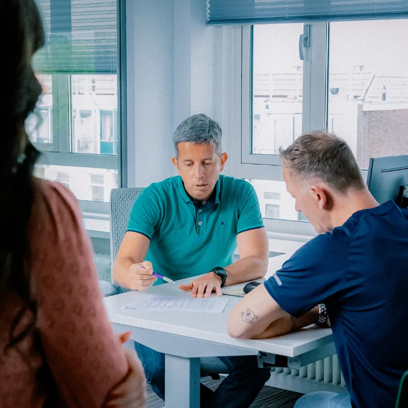 Een behandelaar geeft uitleg aan een cliënt over een formulier aan een bureau. Iemand anders kijkt toe van een afstand.